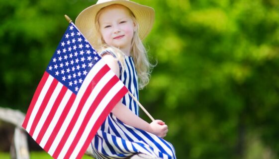 Adorable little girl wearing hat holding american flag outdoors on beautiful summer day